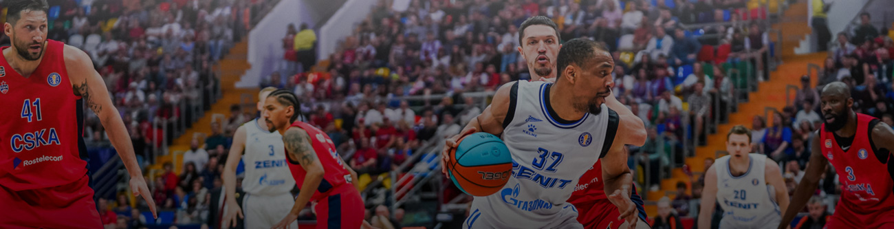 На изображении баскетбольный матч: игрок в белой форме с мячом, окружают игроки в красной и белой форме. Виден трибун с болельщиками на заднем плане