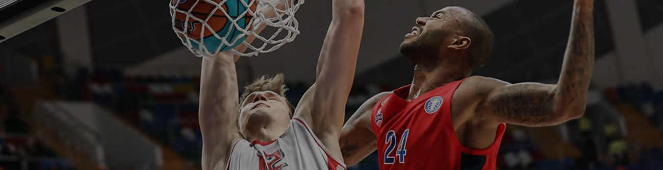 Игрок в белой форме пытается забросить мяч в корзину, тогда как игрок в красной форме пытается защитить кольцо. Они находятся на баскетбольной площадке в крытом спортивном комплексе