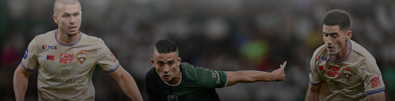 Три футболиста в игровом моменте на поле: игрок в зелёной форме атакует, двое в светлой форме защищаются. Фон размытый, видны зрители на трибунах