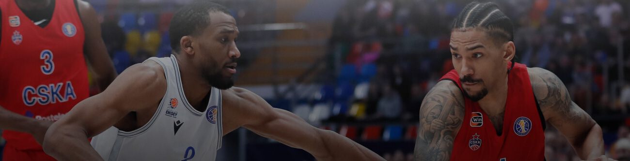 На изображении запечатлен момент баскетбольного матча. Игрок в белой форме пытается пройти защиту игрока в красной форме. На заднем плане видны зрители и элементы спортивного зала